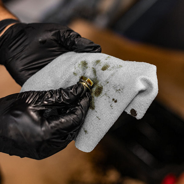 A close up view of a man using a single Rip n Rag multi-purpose microfiber towel from The Rag Company in a grey color to clean oil from an oil pan drain bolt