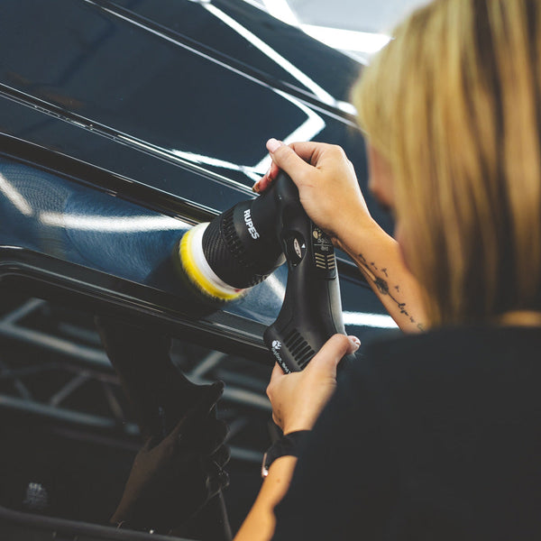 A closeup of a person polishing a black car with a Rupes polisher.